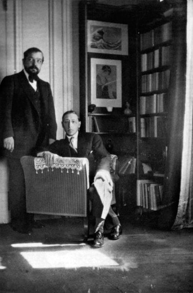 Photograph of Debussy standing next to Stravinsky sitting in a chair inside near a bookshelf