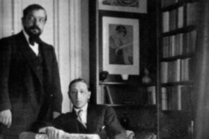 Photograph of Debussy standing next to Stravinsky sitting in a chair