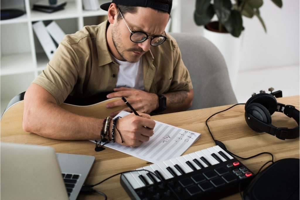 man writing music with a keyboard, laptop, and headphones on a desk